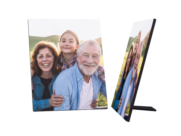 Ein Fotoaufsteller zeigt ein fröhliches Familienbild. Eine lächelnde ältere Frau mit braunen Haaren steht links. Hinter ihr steht ein junges Mädchen mit blonden Haaren in einem karierten Hemd und lächelt ebenfalls. Rechts von ihnen steht ein älterer Mann mit weißen Haaren und Bart, der auch fröhlich in die Kamera schaut und seine Arme um die beiden anderen gelegt hat.