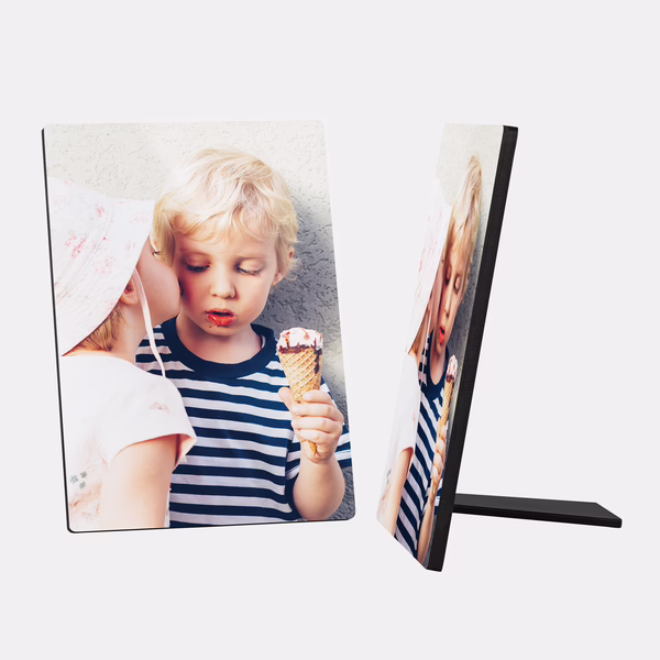 Ein Fotoaufsteller zeigt ein süßes Bild von zwei kleinen Kindern. Ein blondhaariger Junge mit einem blau-weiß gestreiften Shirt hält ein Eis in der Hand und schaut darauf. Ein anderes Kind mit einem hellen Sonnenhut beugt sich zu ihm und scheint ihm einen Kuss auf die Wange zu geben. Der Hintergrund ist hell und schlicht. Rechts daneben ist ein weiterer Fotoaufsteller mit dem gleichen Motiv zu sehen, der auf seiner Stütze steht.